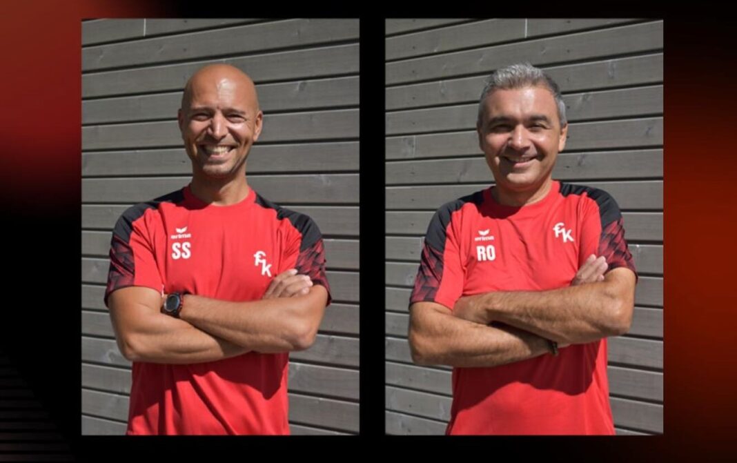 Two men in red sports shirts with black sleeves stand side by side, arms crossed, smiling in front of a gray wooden wall; initials 'SS' and 'RO' appear on their chests.