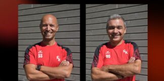Two men in red sports shirts with black sleeves stand side by side, arms crossed, smiling in front of a gray wooden wall; initials 'SS' and 'RO' appear on their chests.