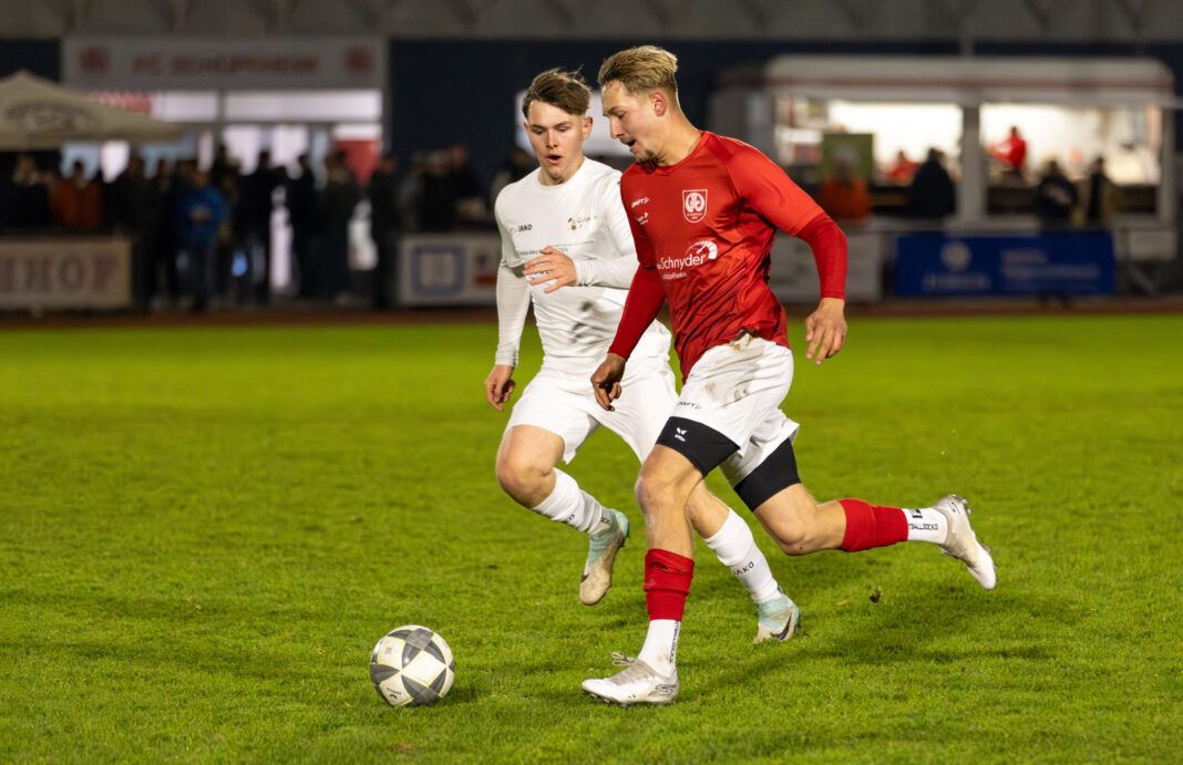 Two soccer players chase a ball on a grassy field during a night match; one wears red, the other white, with spectators in the background at a stadium.