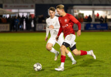 FC Schüpfheim antwortet mit klarem 5:0-Heimsieg Two soccer players chase a ball on a grassy field during a night match; one wears red, the other white, with spectators in the background at a stadium.