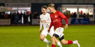 Two soccer players chase a ball on a grassy field during a night match; one wears red, the other white, with spectators in the background at a stadium.