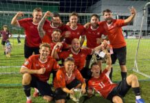 Heiss, heisser, Turnierwochenende FC Gunzwil 2026 Soccer team in red jerseys celebrating with a trophy on a grassy field after a win, group posing inside goal net at night.