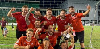 Soccer team in red jerseys celebrating with a trophy on a grassy field after a win, group posing inside goal net at night.