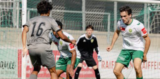 Soccer players contest the ball near the goal as a goalkeeper watches from the netted area in the background.