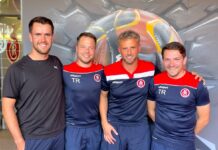 Trainerstaff der 1. Mannschaft mit viel FC Rothenburg ergänzt Four men in navy and red training outfits pose together, smiling in front of an urban mural.”,
