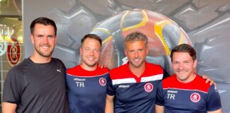 Four men in navy and red training outfits pose together, smiling in front of an urban mural.”,