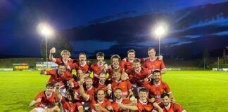 A soccer team in red uniforms posing on a grass field at night with a trophy, celebrating their win.