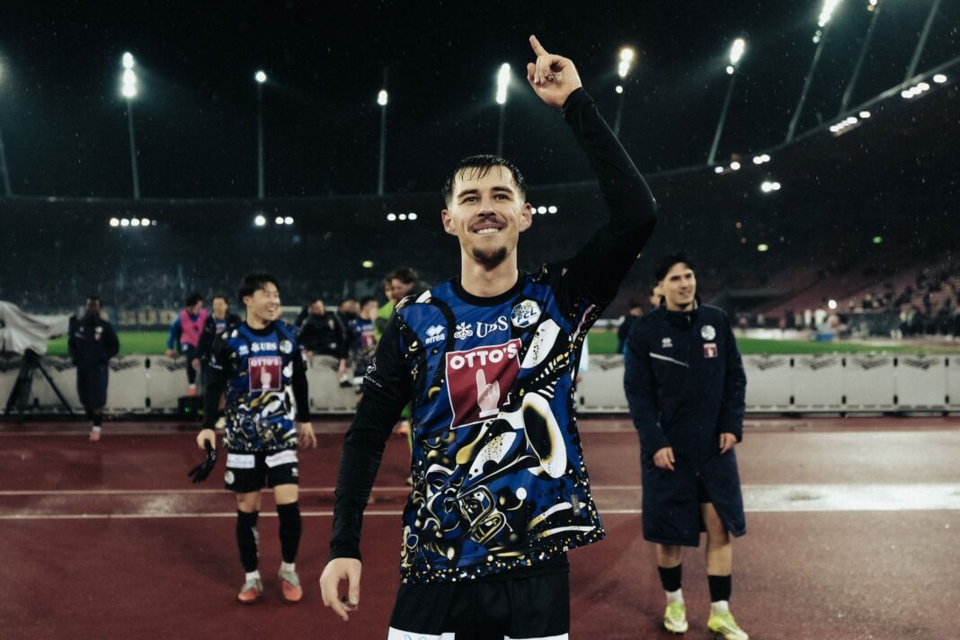adrian-bajrami-transfer Athlete on a track field raises a finger in victory, lights glow in the rainy stadium behind him.