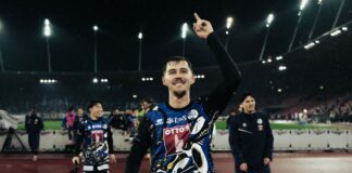 Athlete on a track field raises a finger in victory, lights glow in the rainy stadium behind him.