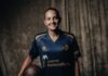 FCL: AWSL-Team verpflichtet Athanasia Moraitou Smiling woman in a blue sports jersey holding a brown football, standing against a dark textured backdrop.