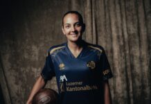 FCL: AWSL-Team verpflichtet Athanasia Moraitou Smiling woman in a blue sports jersey holding a brown football, standing against a dark textured backdrop.