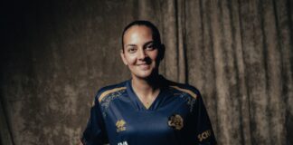 Smiling woman in a blue sports jersey holding a brown football, standing against a dark textured backdrop.