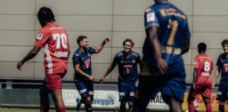 Blue soccer players celebrate a goal on a sunny field, central player smiling as teammates approach from behind red-clad opponents.