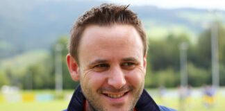 Smiling man in a blue Adidas jacket outdoors on a sports field.