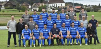 Group of soccer players in blue jerseys and staff posing on a grassy field with houses in the background.