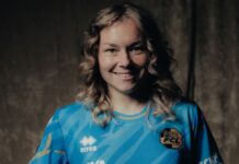 FCL: Laura Vogt stösst zum AWSL-Team Smiling woman in a blue sports jersey with logos, posed against a dark backdrop.