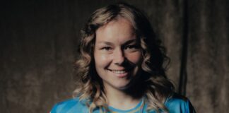Smiling woman in a blue sports jersey with logos, posed against a dark backdrop.