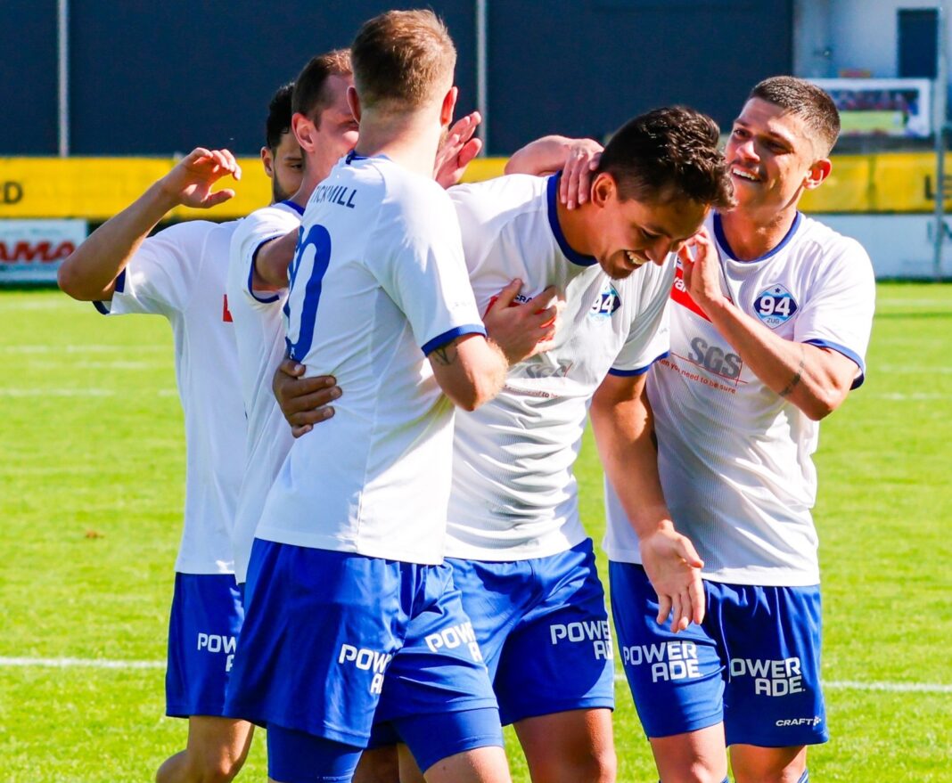 Soccer players in white jerseys celebrate a goal on a sunny green field.