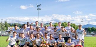 Group of soccer players in white jerseys posing for a victory photo on a sunny field, holding a trophy.