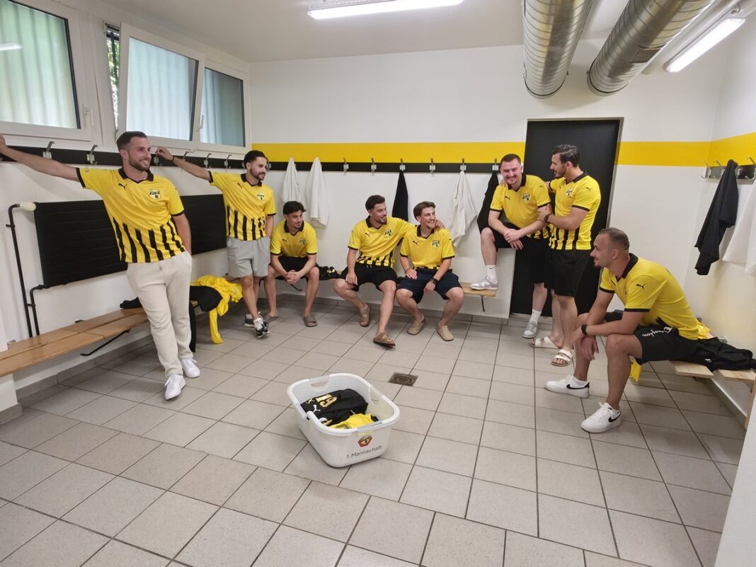 20260624_192749 Group of men in yellow and black jerseys gather in a locker room, some sitting on benches and others leaning against the wall.