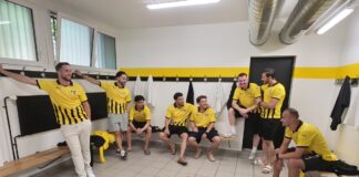 Group of men in yellow and black jerseys gather in a locker room, some sitting on benches and others leaning against the wall.