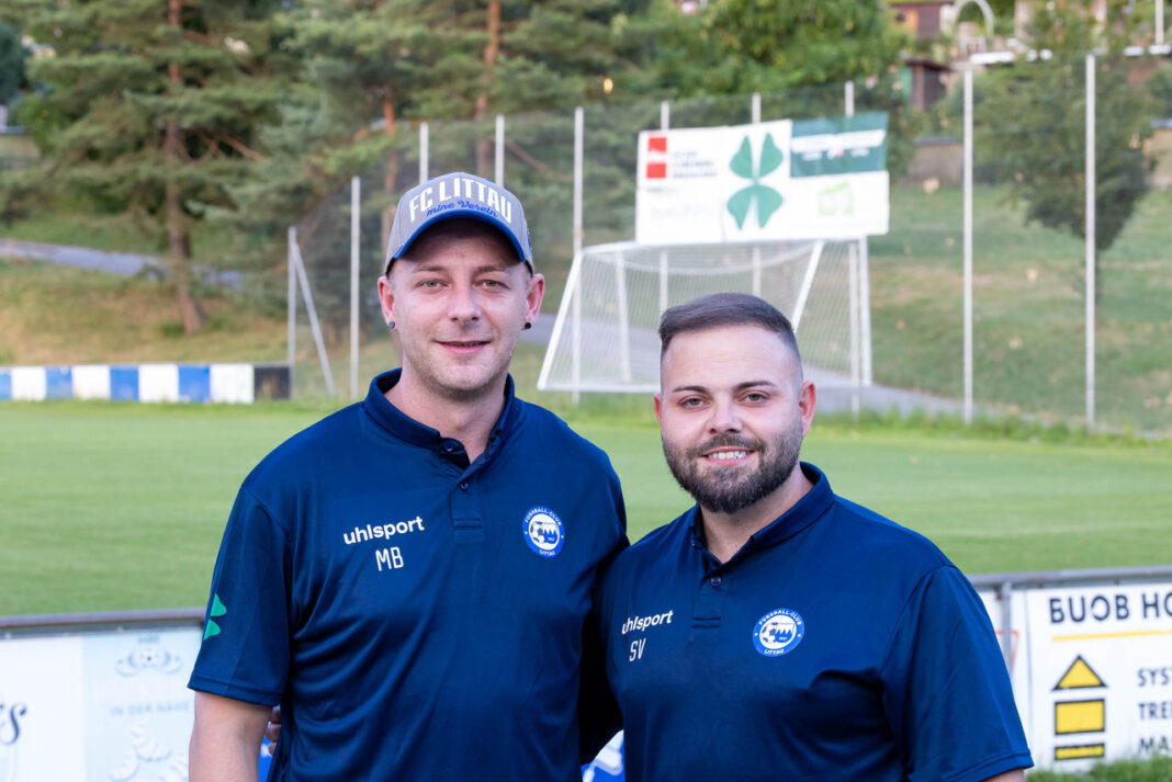 2026_07_06_45895 Two coaches in matching navy polo shirts stand together on a grassy soccer field, smiling at the camera with a goal and banners in the background.