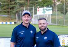 FC Littau verpflichtet neuen Assistenz- und Torhütertrainer Two coaches in matching navy polo shirts stand together on a grassy soccer field, smiling at the camera with a goal and banners in the background.