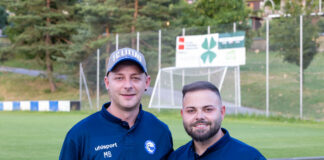Two coaches in matching navy polo shirts stand together on a grassy soccer field, smiling at the camera with a goal and banners in the background.