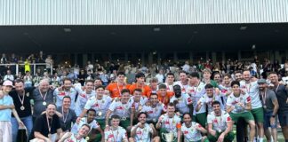 Group of soccer players and staff posing on the field with medals and a trophy after a win, stadium stands in the background.