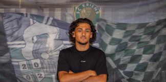 Young man with arms crossed wearing a black t-shirt, standing in front of a soccer team banner behind him.