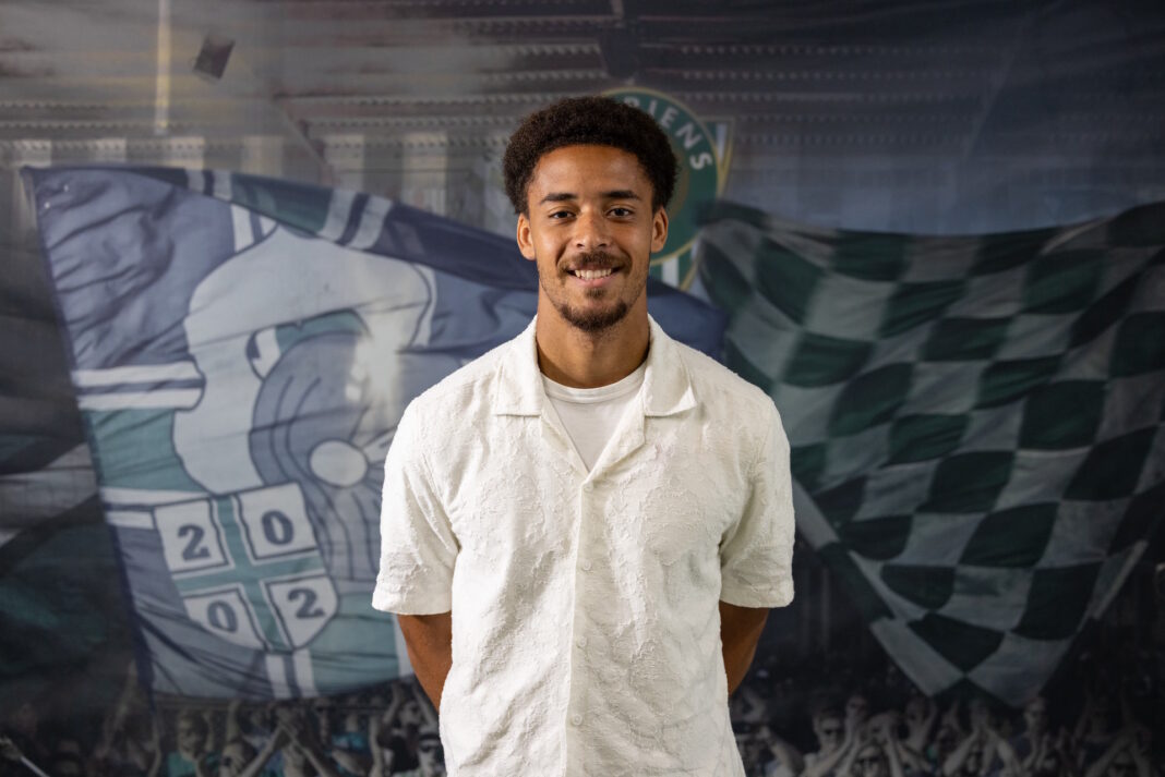 364A2228 Smiling man with short curly hair wearing a white textured shirt, posed in front of a sports-themed backdrop with flags and crowd imagery.
