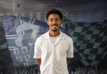 Marvin Akahomen wechselt vom FC Basel zum SC Kriens Smiling man with short curly hair wearing a white textured shirt, posed in front of a sports-themed backdrop with flags and crowd imagery.