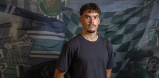 Portrait of a young man in a dark T-shirt standing in front of a green-and-white sports banner backdrop.