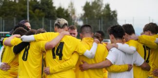 Soccer team in yellow uniforms huddles with arms around each other on a grassy field, seen from behind.