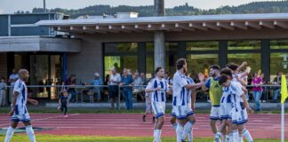 Soccer players in blue-and-white striped kits celebrate together near the track while a few spectators watch from behind a railing.