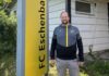 Andreas Müller: Verwurzelt in Eschenbach, engagiert für die Zukunft Man standing outdoors beside a tall yellow sign with vertical black lettering, wearing a gray and black jacket with yellow accents, in front of a light-colored house.
