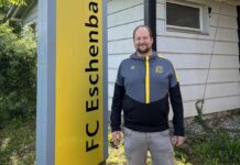 Andreas Müller: Verwurzelt in Eschenbach, engagiert für die Zukunft Man standing outdoors beside a tall yellow sign with vertical black lettering, wearing a gray and black jacket with yellow accents, in front of a light-colored house.