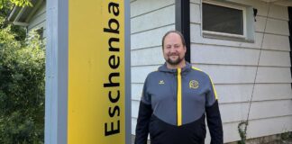 Man standing outdoors beside a tall yellow sign with vertical black lettering, wearing a gray and black jacket with yellow accents, in front of a light-colored house.