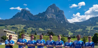 Group of athletes in matching blue jerseys posing for a team photo on a track with green fields and mountains in the background.
