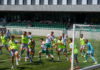 Kriens testete gegen Schötz und YF Juventus Soccer players in neon vests collide near the goal while a goalkeeper in turquoise watches from the right, in a stadium with green seats and spectators in the background.