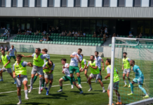 Kriens testete gegen Schötz und YF Juventus Soccer players in neon vests collide near the goal while a goalkeeper in turquoise watches from the right, in a stadium with green seats and spectators in the background.