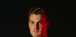 Portrait of a man in a white sports jersey with a crest, facing the camera against a dark background.