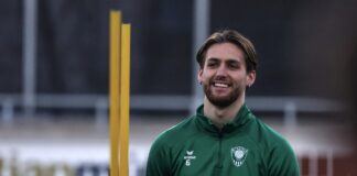 Smiling man in a green training jacket with number 6 stands on a sports field, orange training posts in the background.