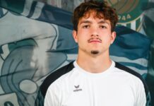 Kevin Tröndle: Vom Basler Nachwuchs ins Kleinfeld Portrait of a young man with brown hair and a light goatee, wearing a white and black athletic shirt, standing before a blue mural backdrop.