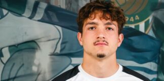Portrait of a young man with brown hair and a light goatee, wearing a white and black athletic shirt, standing before a blue mural backdrop.