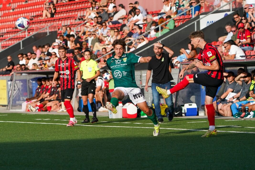 Soccer match action: green player and red-black player contest a high ball near the sideline with a crowd in the stands behind them.