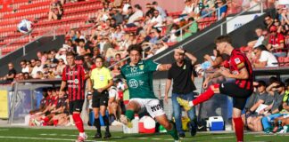 Soccer match action: green player and red-black player contest a high ball near the sideline with a crowd in the stands behind them.
