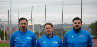 Three men in matching blue athletic jackets stand side by side on a football training field, looking at the camera.