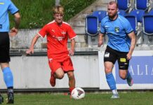 Cupfieber im IFV: Die erste Runde verspricht Spannung und Überraschungen Red team player dribbles the ball while a blue-team opponent pursues on a grassy soccer field near empty blue seats in the stands.