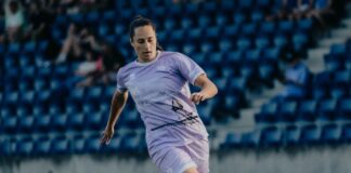Female soccer player in a light purple uniform dribbles a ball on a grass field, with blue stadium seats in the background.